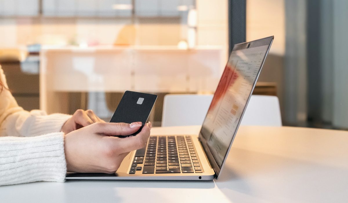 Pessoa segurando um cartão preto em frente a um notebook aberto sobre a mesa