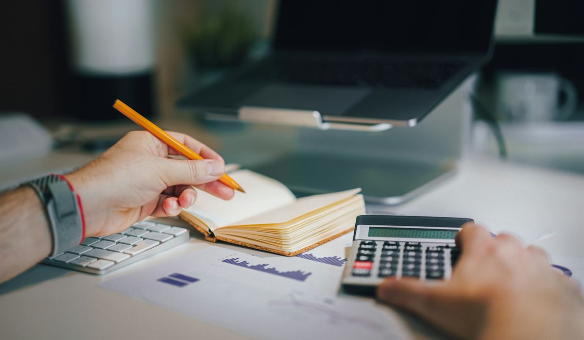 Pessoa fazendo contas com calculadora e anotando em um caderno sobre a mesa